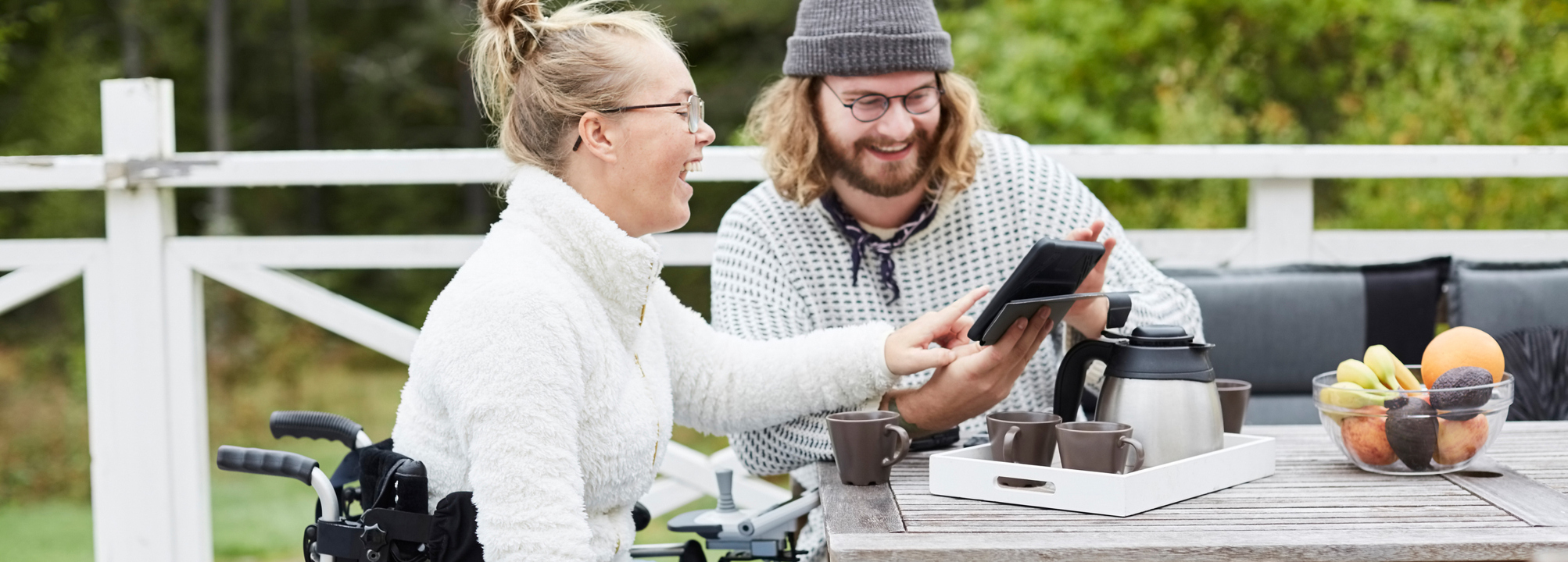 Manlig vårdgivare och kvinna i rullstol sitter vid bord på altan med mobiltelefon.